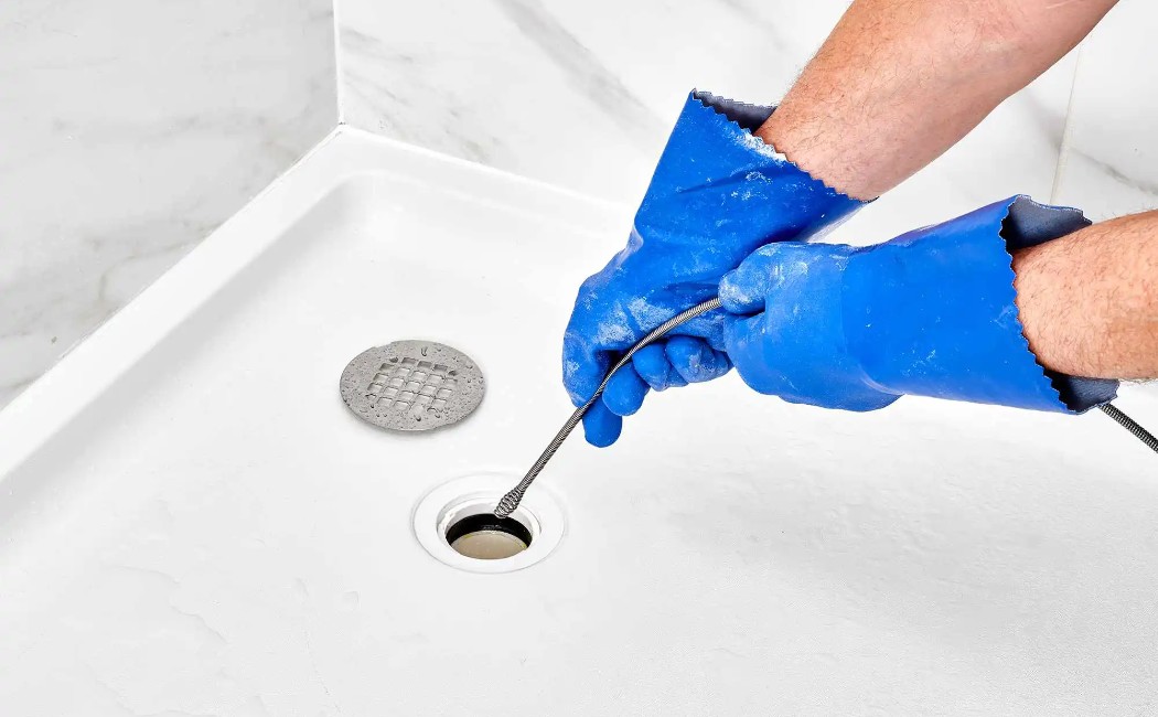 Technician wearing protective gloves using a drain snake to clear a clogged shower drain, highlighting professional Drain Cleaning Services for smooth water flow.