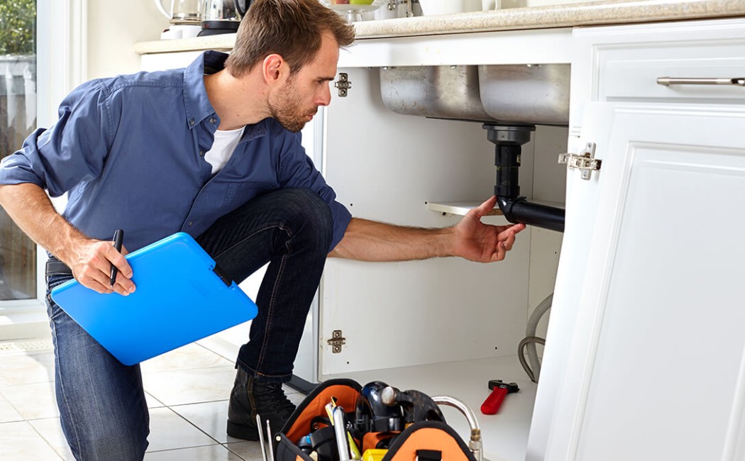 Technician performing a plumbing inspection while checking the kitchen sink drain pipes inside a cabinet.