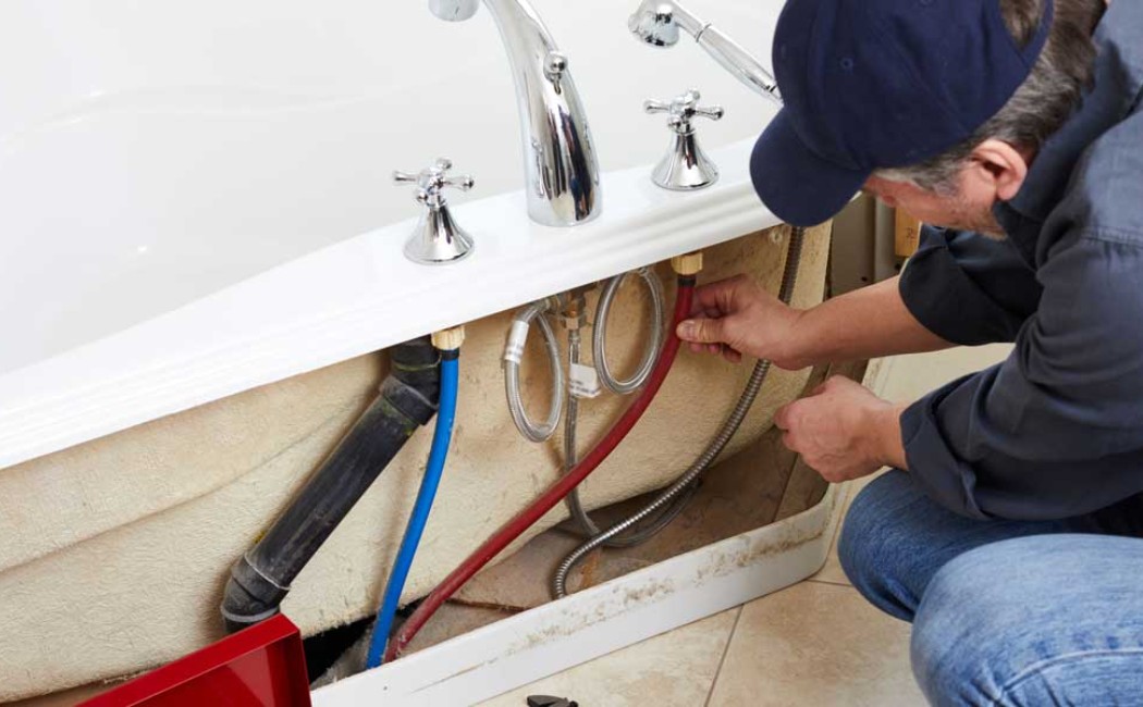 Plumber providing Bathroom Plumbing Services by connecting water supply lines under a bathtub and inspecting pipes for proper installation and leaks.