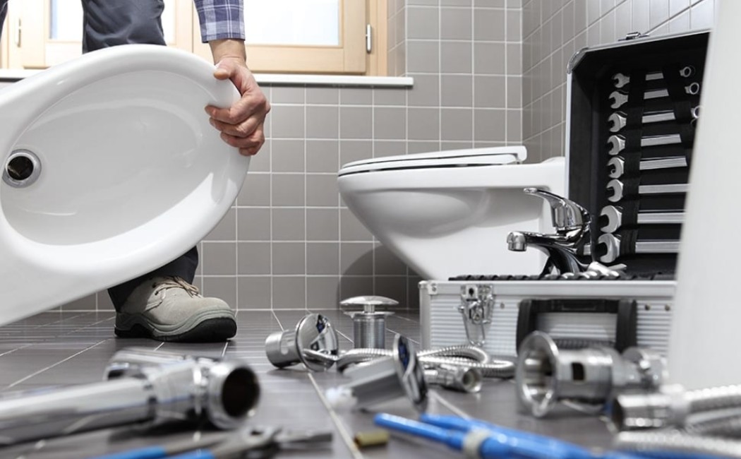 Technician performing Bathroom Plumbing Services while installing a sink basin with plumbing tools and fittings spread across a tiled bathroom floor.