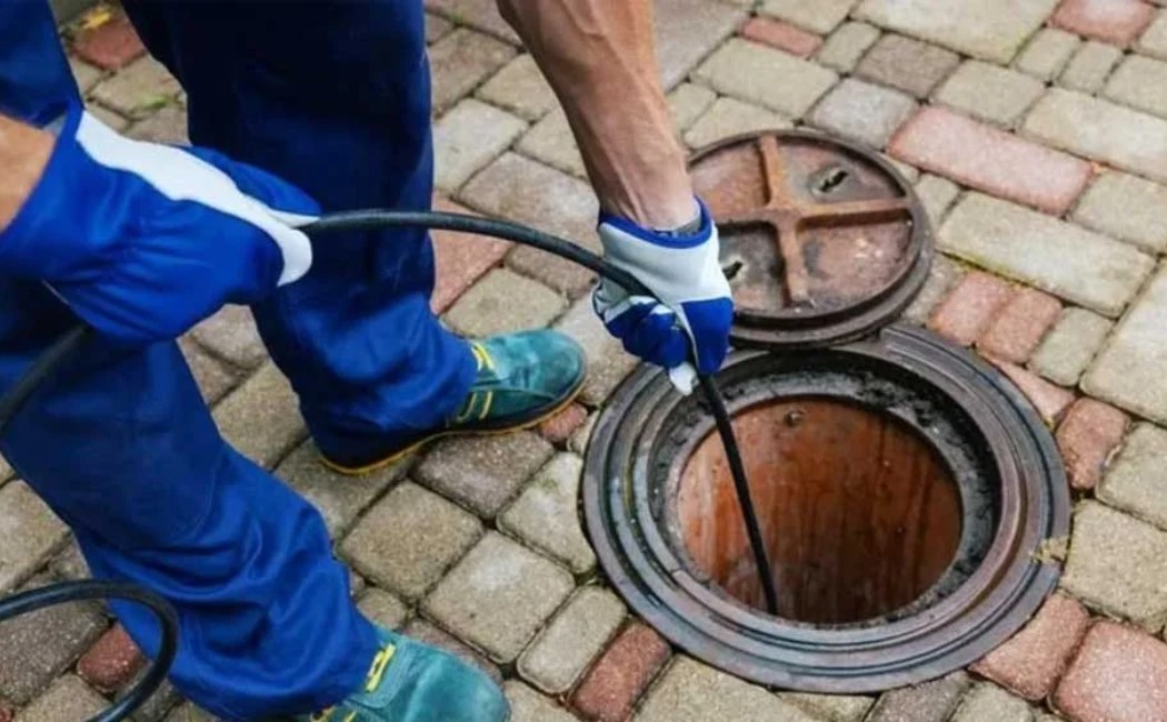 Technician performing drain cleaning by inserting a hose into an outdoor manhole on a paved surface.
