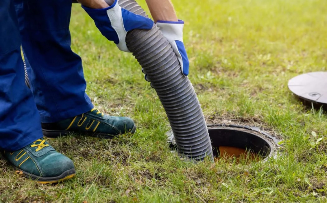 Worker performing drain cleaning by inserting a large hose into an outdoor sewer opening on a grassy area.