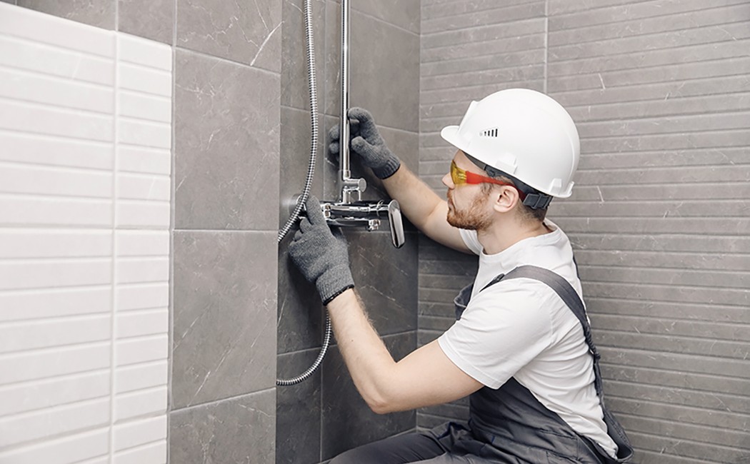 Bathroom Shower Fixture Installation Expert Worker wearing a hard hat and safety glasses installing a shower fixture in a tiled bathroom.