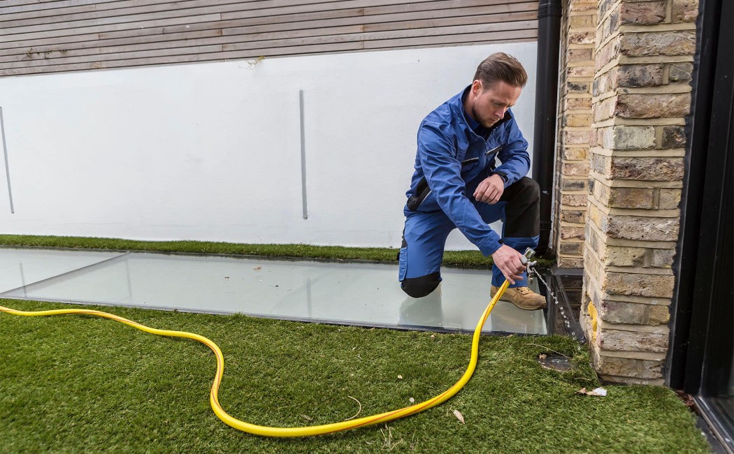 Plumber performing Leak Detection Services by inspecting an outdoor water tap and drainage area near a residential building.
