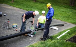 Roadway Sewer Pipe Installation and Repair Work. Utility workers performing Sewer Repair Services by cutting and installing large underground pipes on a residential roadway with professional equipment and safety gear.