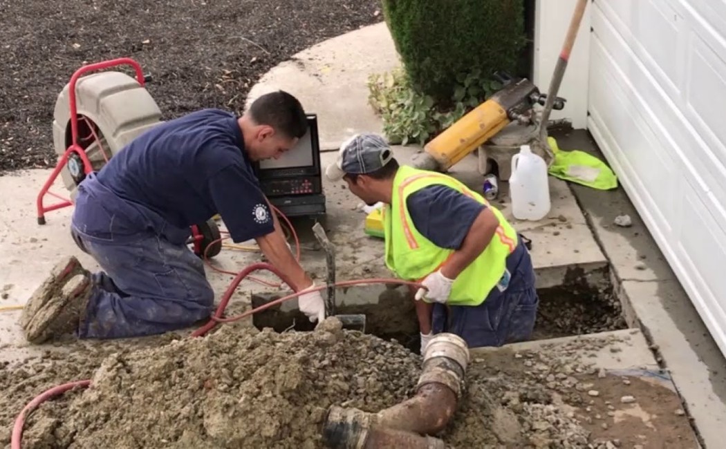 Workers performing Sewer Installation by excavating near a residential driveway and inspecting underground piping with specialized equipment.